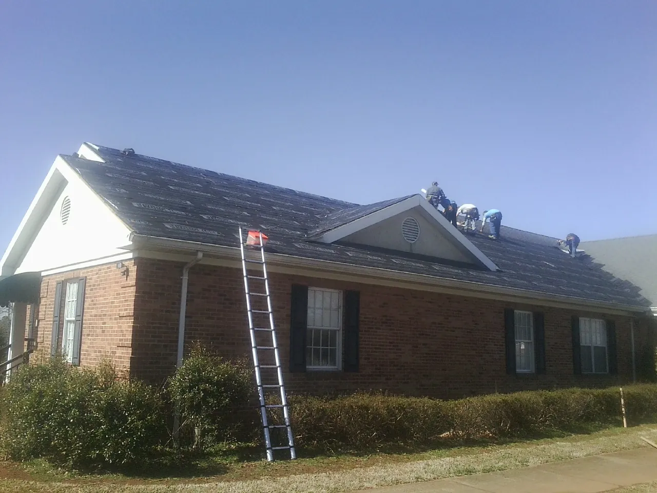 Crew tearing shingles off a brick ranch roof with a ladder set — Emergency Roof Repair work by our crew in Little Falls